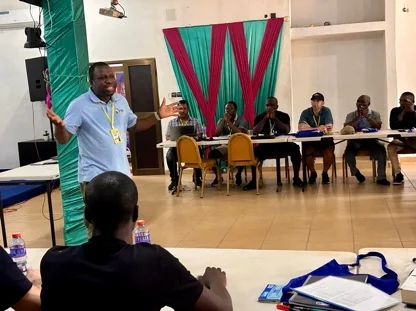 A facilitator wearing a lanyard stands and gestures while addressing seated participants at the African Disciple-Making Movement Leadership Challenge Summit in Lomé, Togo, with colorful draped fabric in the background