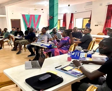 Summit participants seated at tables with laptops and notebooks during a working session at the DMM Leadership Challenge Summit in Lomé, Togo, with colorful green and red fabric decorations on the wall behind them
