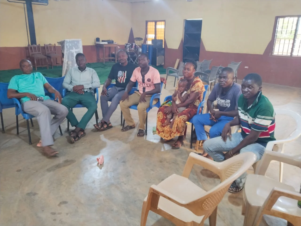 A small group of young people seated on blue plastic chairs in a circle inside a church hall with a drum set and speakers on stage, during a session on faith and business led by Samson Abukari in Ghana