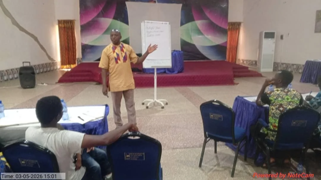 A facilitator in a traditional Ghanaian shirt stands and gestures beside a flip chart while teaching church planters seated in blue upholstered chairs inside a conference hall with a decorated stage