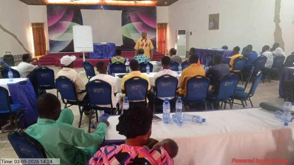 Wide view of a church planter training session in a conference hall in Ghana, with approximately twenty participants seated at cloth-covered tables facing a standing facilitator, flip chart and stage backdrop visible behind him
