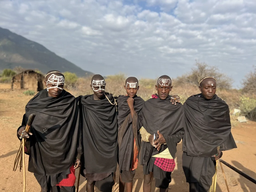 Five young Maasai men wearing black cloaks and traditional white face paint pose together with arms around each other, with mountains and dry savanna landscape in the background in the Nkewua community