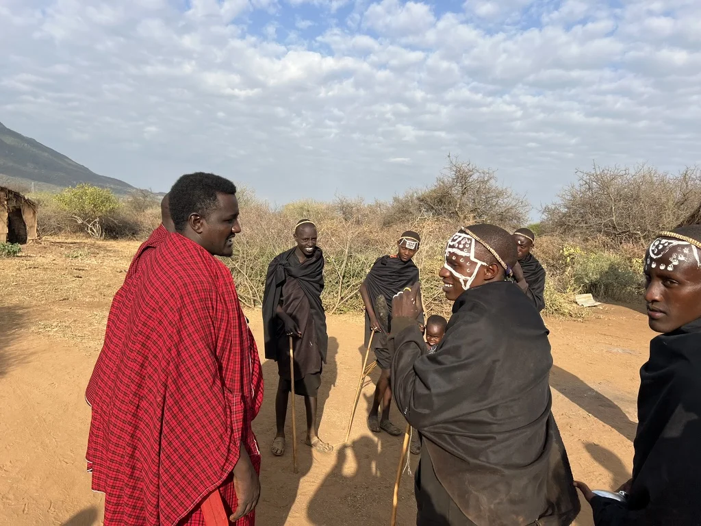 A man in a red Maasai shuka speaks with a group of young men in black cloaks with white face paint, standing on dry red earth with mountains and scrubland behind them
