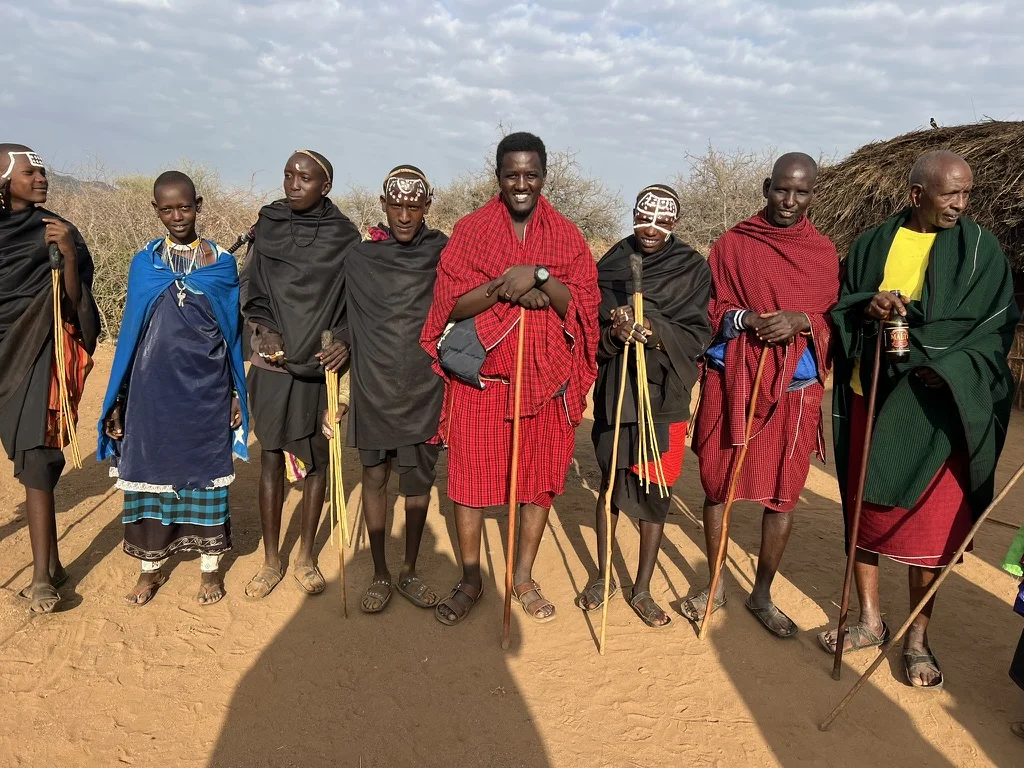 A group of Maasai men and youth of various ages standing in a line, some wearing red shukas and others in black cloaks with white face paint, holding walking sticks, with a thatched dwelling visible behind them