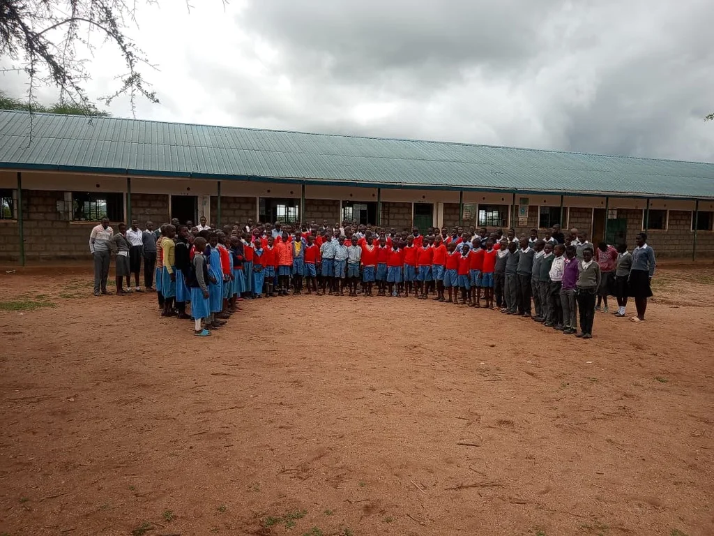 A large group of students wearing red sweaters and blue skirts or shorts standing in a U-shaped formation in the schoolyard in front of a modern concrete-block school building with a green metal roof — Karero Primary School in 2026