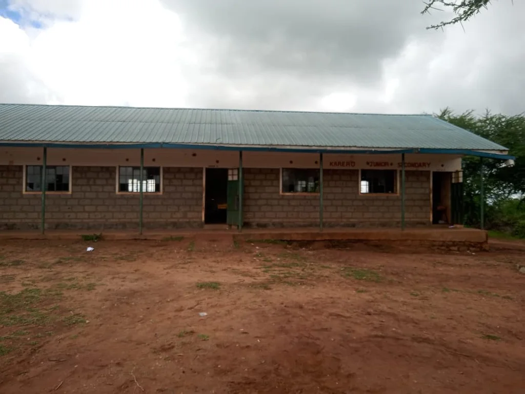 Exterior view of the modern Karero Primary School building in 2026, a concrete-block structure with a green corrugated metal roof, covered veranda, and windows, set on red earth with overcast skies