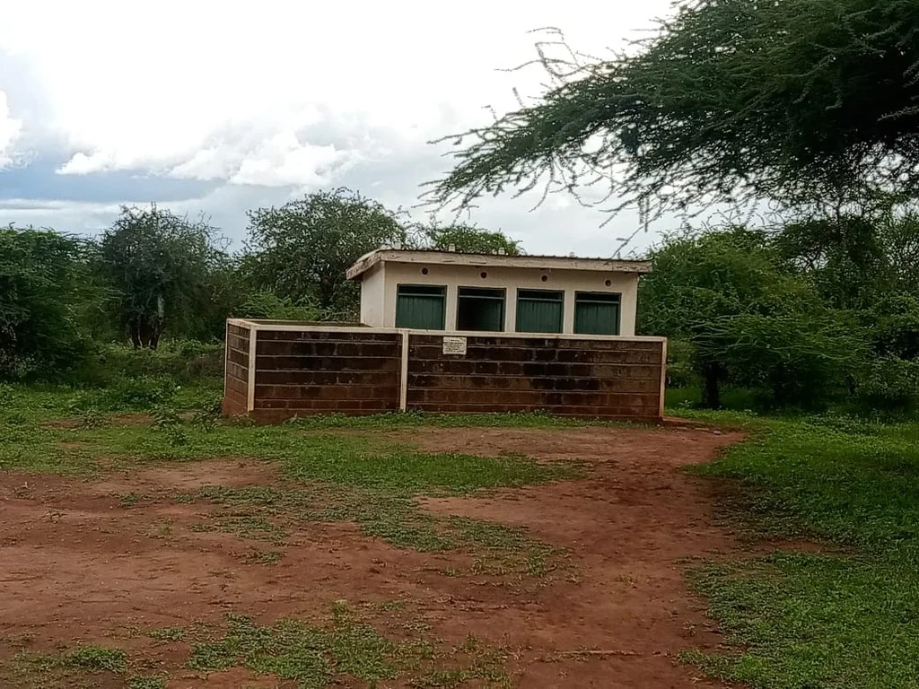 A small concrete latrine building with green doors and a brick perimeter wall, surrounded by acacia trees on red soil — the new sanitation facilities at Karero Primary School