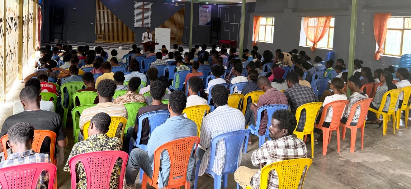 A large group of young people seated on colourful plastic chairs in a church hall, listening to a speaker at the front