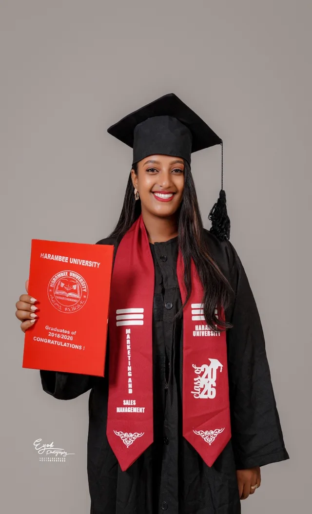 Yanet in a graduation cap and gown with a red Marketing and Sales Management stole, holding a red Harambee University graduation folder