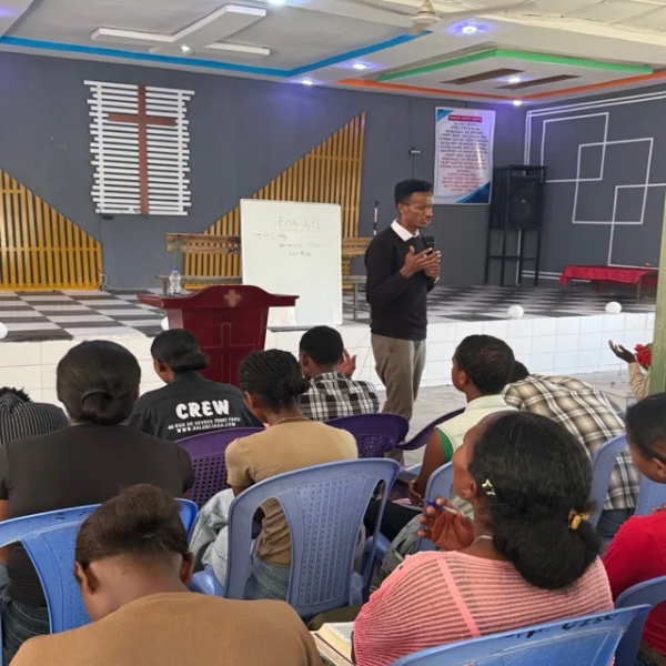 A speaker with a microphone in front of a whiteboard as young people seated in blue chairs listen and take notes