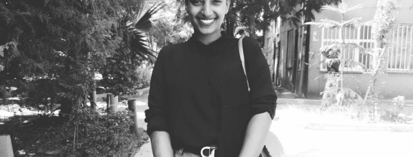 Yanet smiling outdoors in a black-and-white photo, standing on a tree-lined path in front of a building