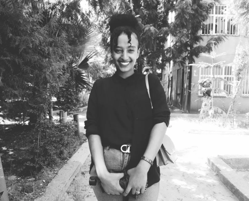 Yanet smiling outdoors in a black-and-white photo, standing on a tree-lined path in front of a building