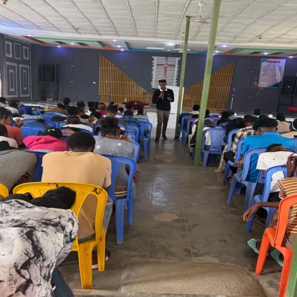 A speaker standing in the aisle with a microphone, teaching young people seated on plastic chairs in a church hall