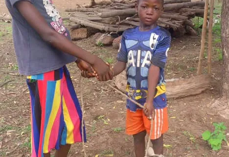 A girl and a young boy shake hands in a village yard as the boy holds a rope tied to a small grey and white goat, with thatched-roof huts and a tree behind them