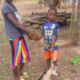 A girl and a young boy shake hands in a village yard as the boy holds a rope tied to a small grey and white goat, with thatched-roof huts and a tree behind them