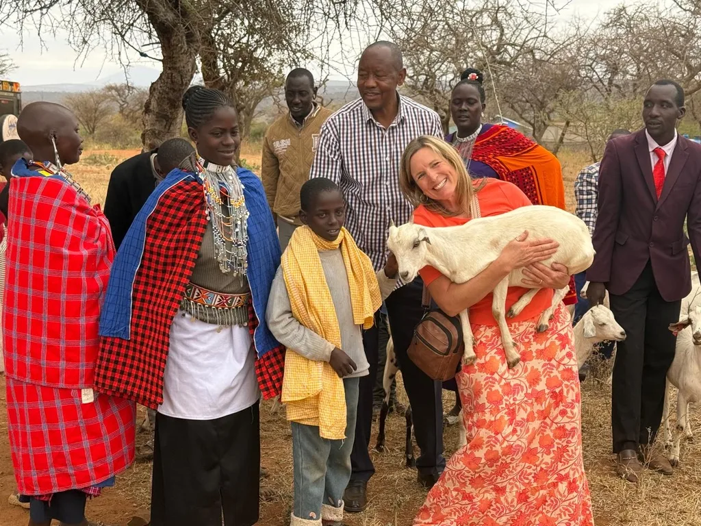 A smiling visitor in an orange dress holding a white goat, standing with children, guardians and church leaders outdoors