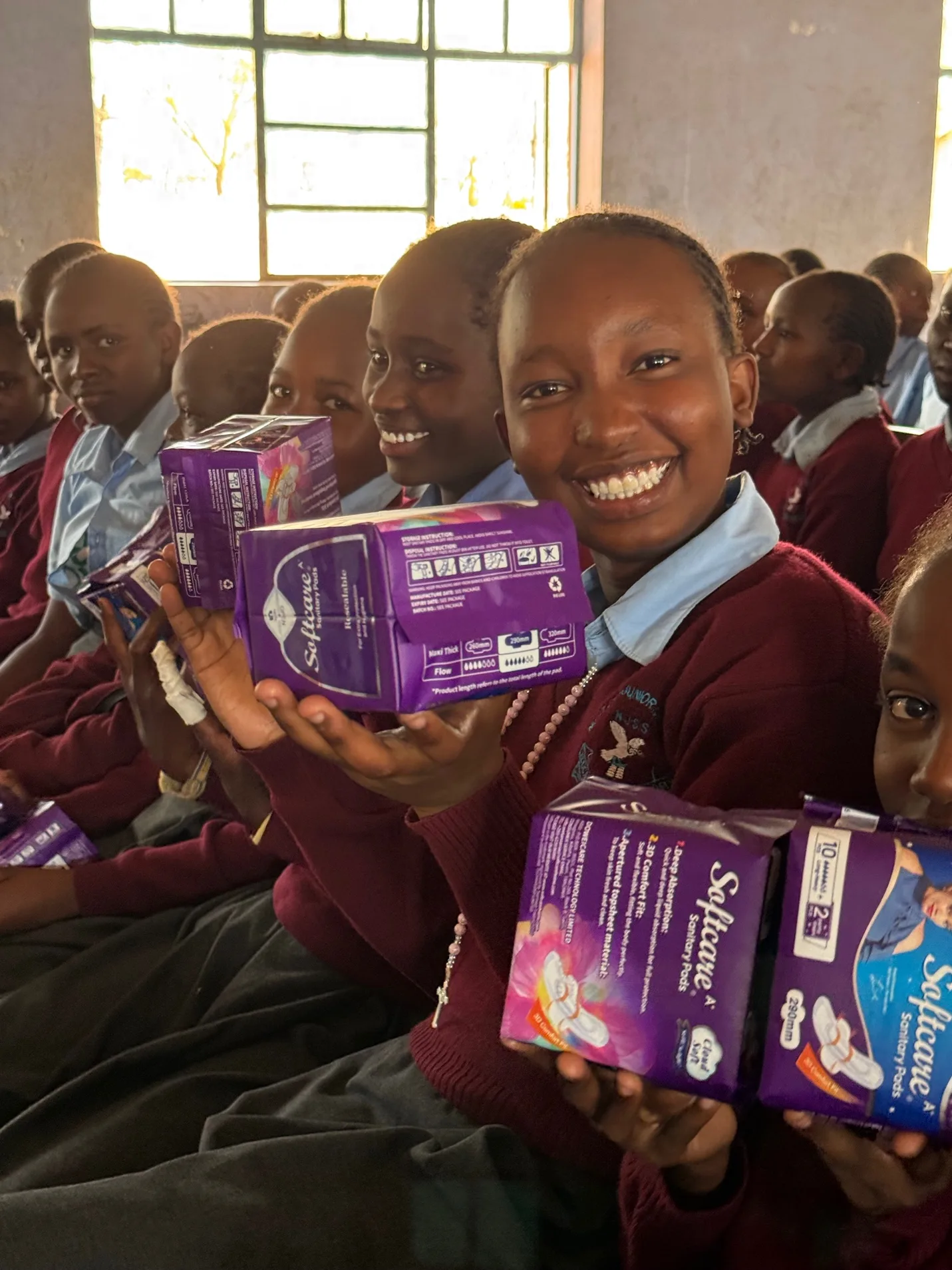 Smiling schoolgirls in maroon uniforms holding up packets of Softcare sanitary pads close to the camera