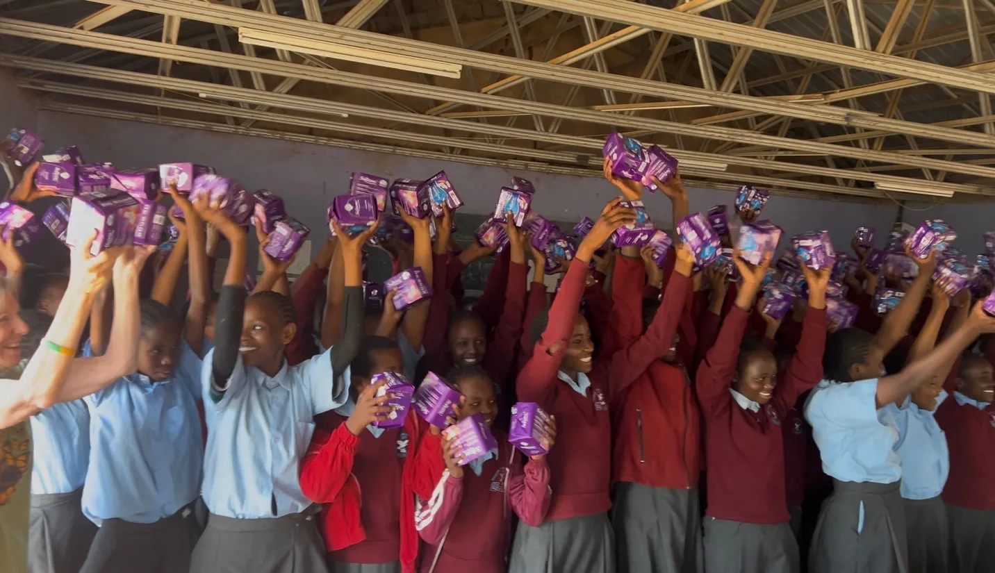 Schoolgirls in maroon and blue uniforms raising packets of dignity pads above their heads in celebration inside a classroom