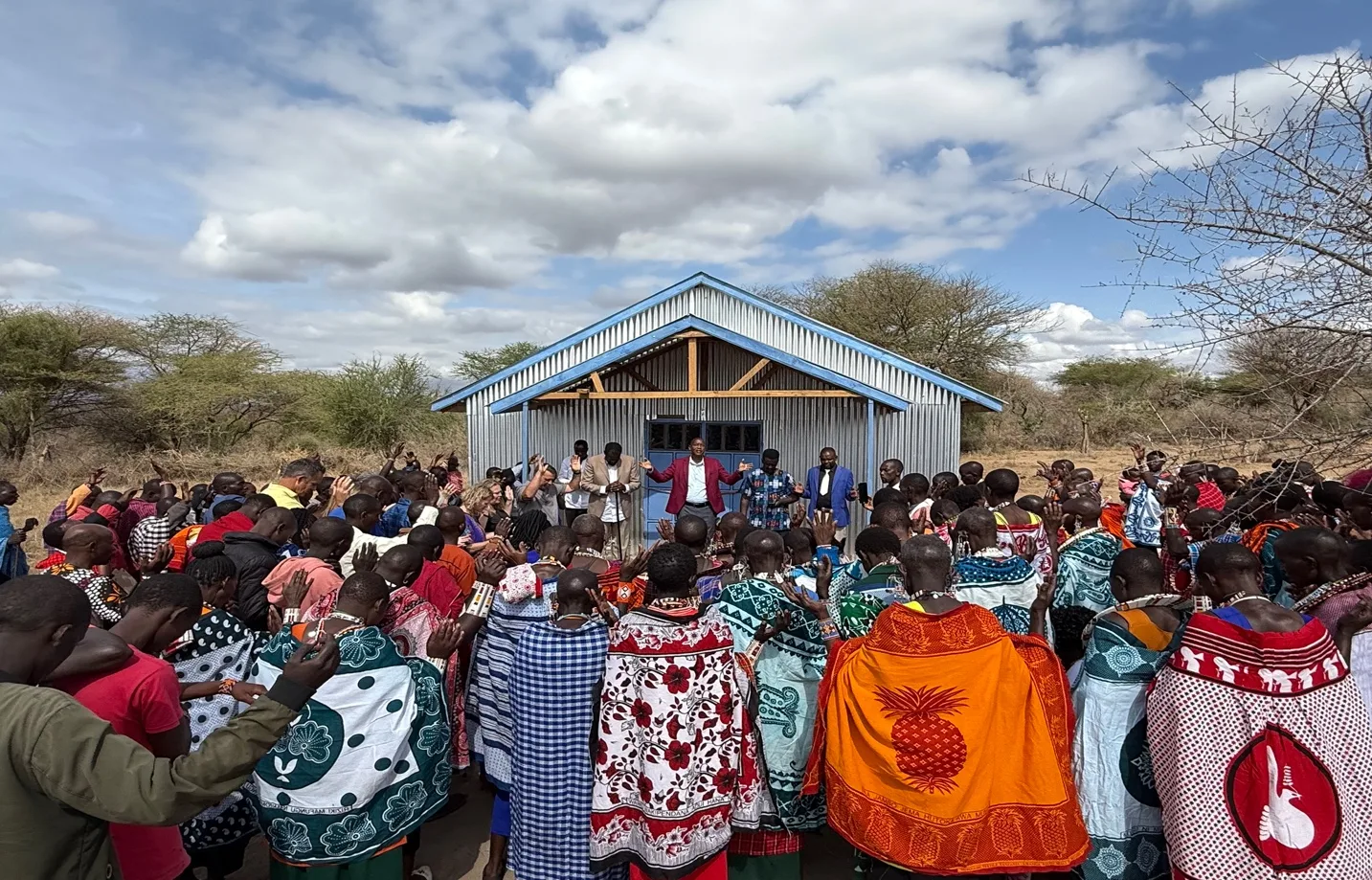 A large crowd of community members in colourful traditional dress gathered in worship outside the new Indoinyo church building under a cloudy sky