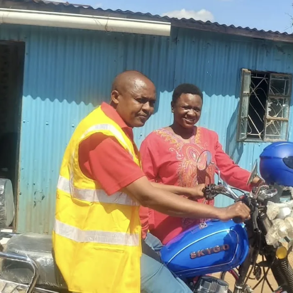 A pastor in a yellow safety vest and a woman in a red patterned top sitting on a new blue motorbike outside a blue corrugated iron building