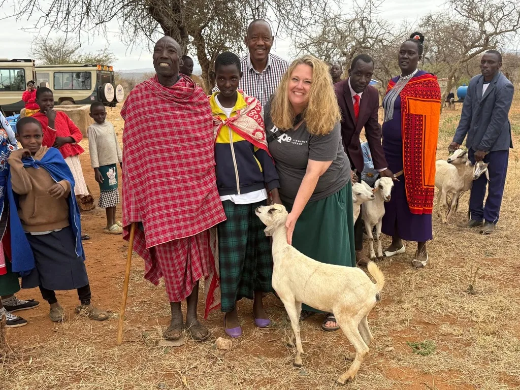 A visitor in a grey T-shirt standing with children, guardians and church leaders around a white goat in a dry, open field