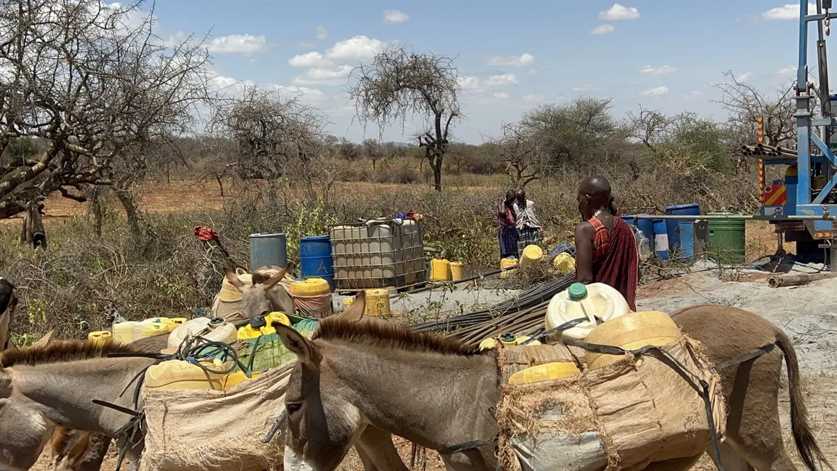 Donkeys loaded with water containers standing near the borehole site, with jerrycans and drilling equipment in the background