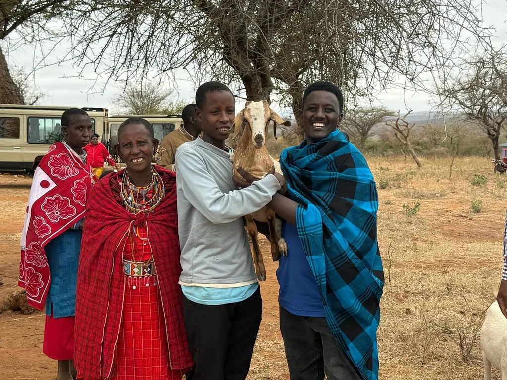 Two young people holding a small goat, standing with women in red Maasai dress in a dry field with safari vehicles behind
