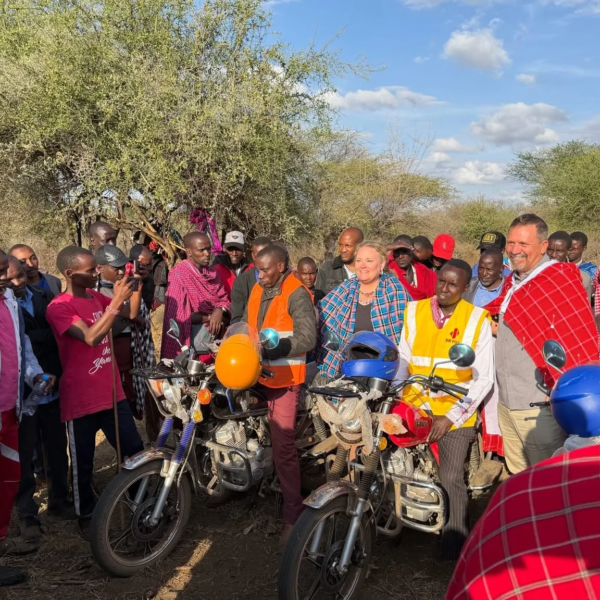 A crowd gathered outdoors among acacia trees around new motorbikes as pastors receive them, with visiting guests standing among community members