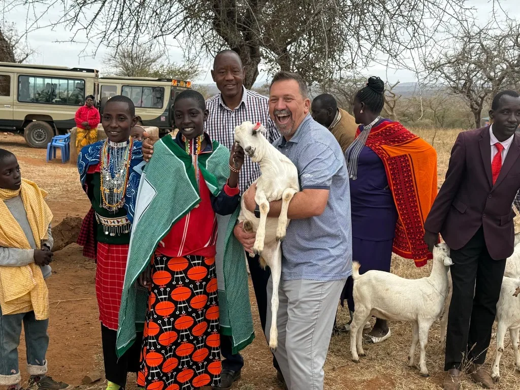 A laughing visitor holding a white goat beside children and community members in traditional dress, with more goats and safari vehicles nearby