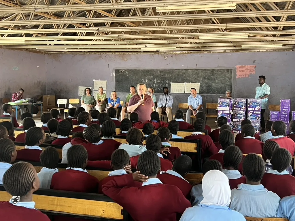 Rows of schoolgirls seated at wooden desks in a classroom listening to a visiting speaker with a microphone, with boxes of dignity pads at the front
