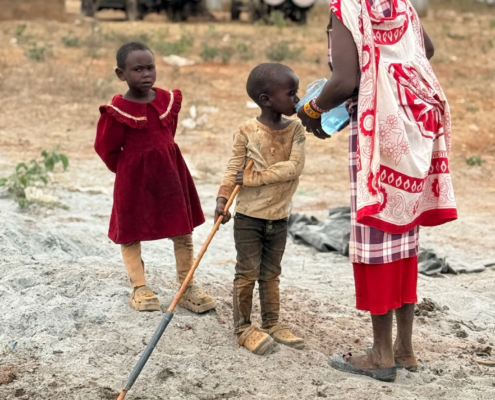A mother in a red shuka gives water from a bottle to a small boy while a young girl in a red dress stands beside them near the borehole