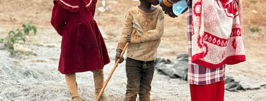 A mother in a red shuka gives water from a bottle to a small boy while a young girl in a red dress stands beside them near the borehole