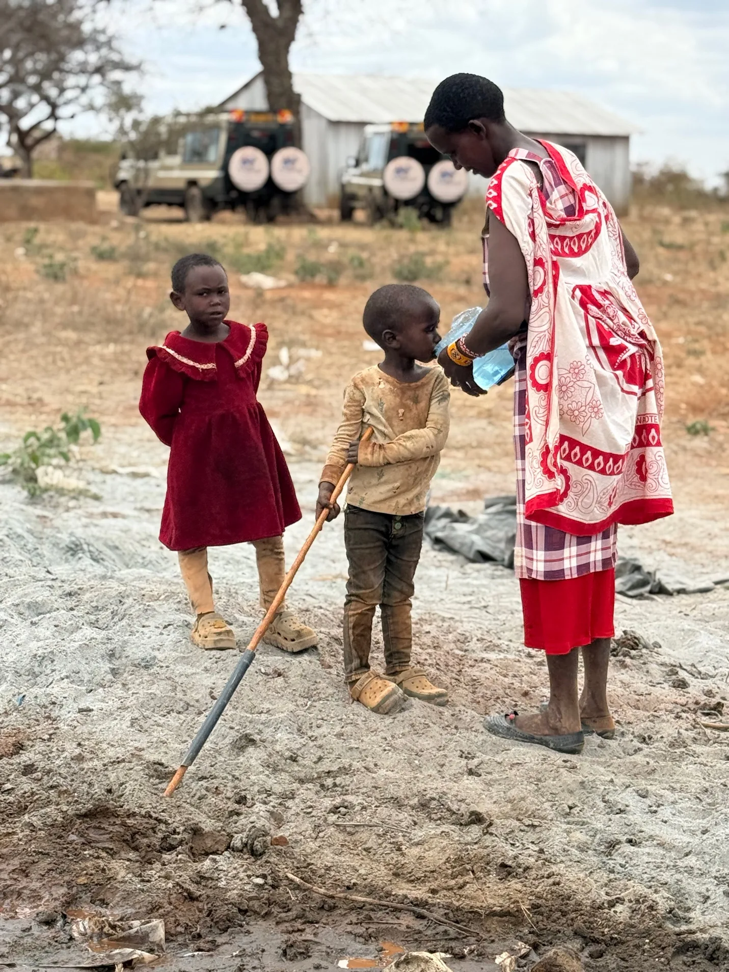 A mother in a red shuka gives water from a bottle to a small boy while a young girl in a red dress stands beside them near the borehole